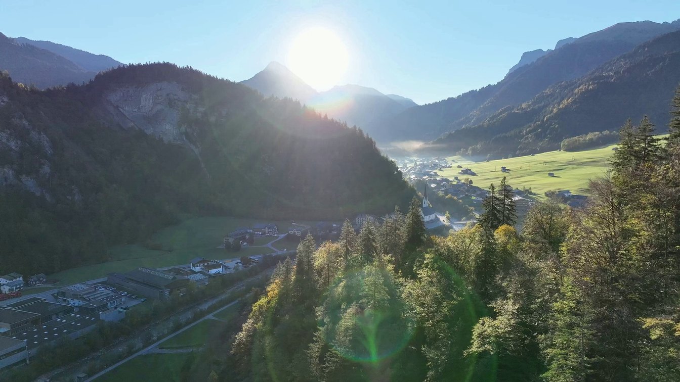 Drohnenflug am Morgen über ein Tal im Bregenzerwald mit Wald, Wiesen, Dorf und Kirche, gegen die Sonne